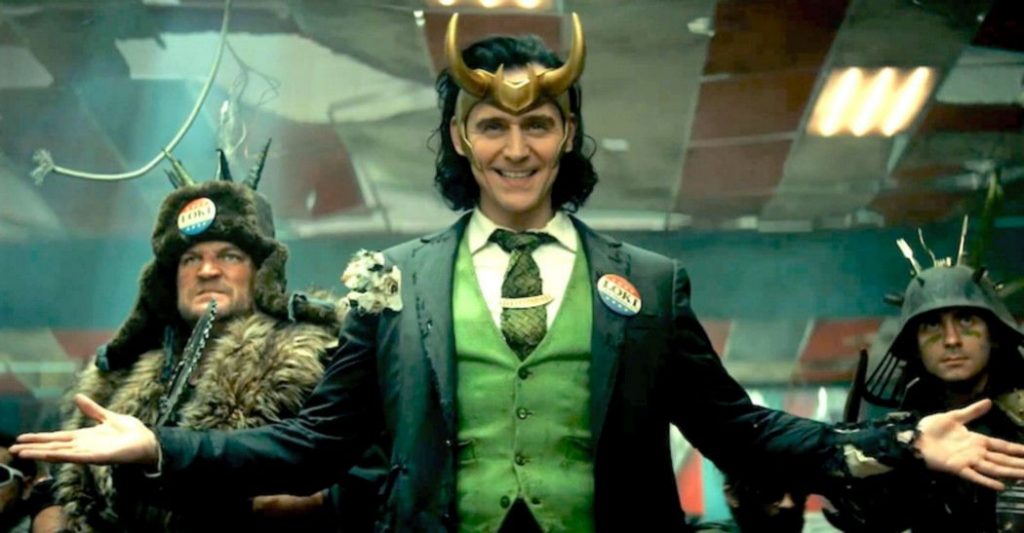 Loki smiles, arms out-stretched, flanked by two men.